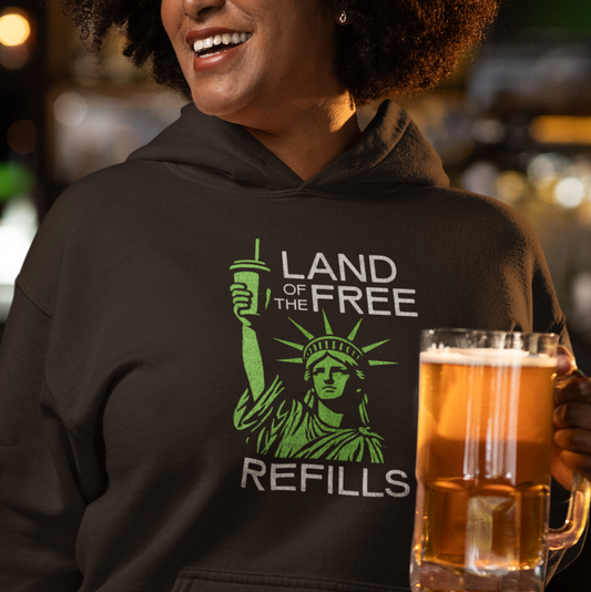Person wearing a hoodie with 'Land of the Free Refills' design, holding a beer mug in a casual setting.