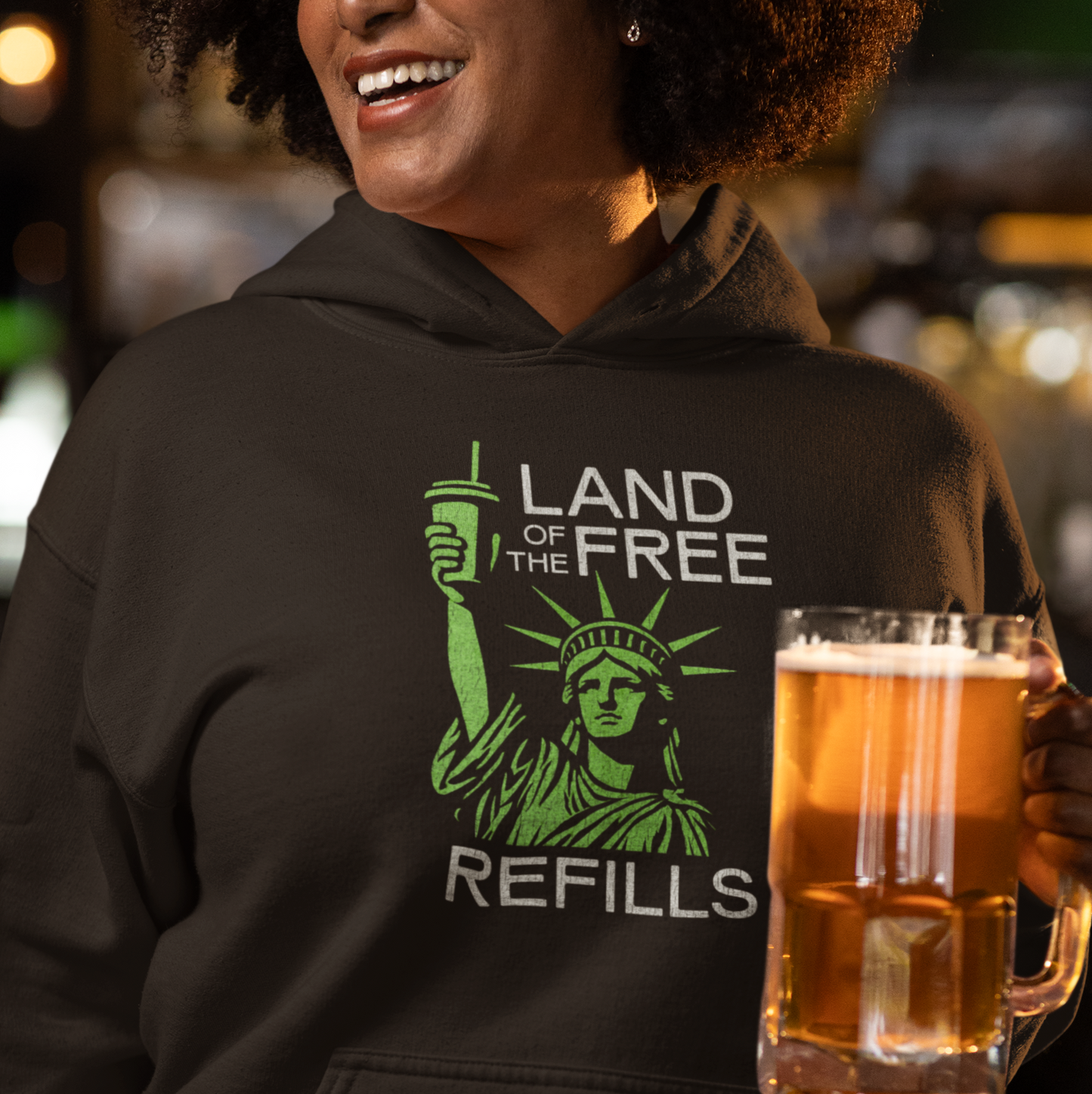 Person wearing a hoodie with 'Land of the Free Refills' design, holding a beer mug in a casual setting.