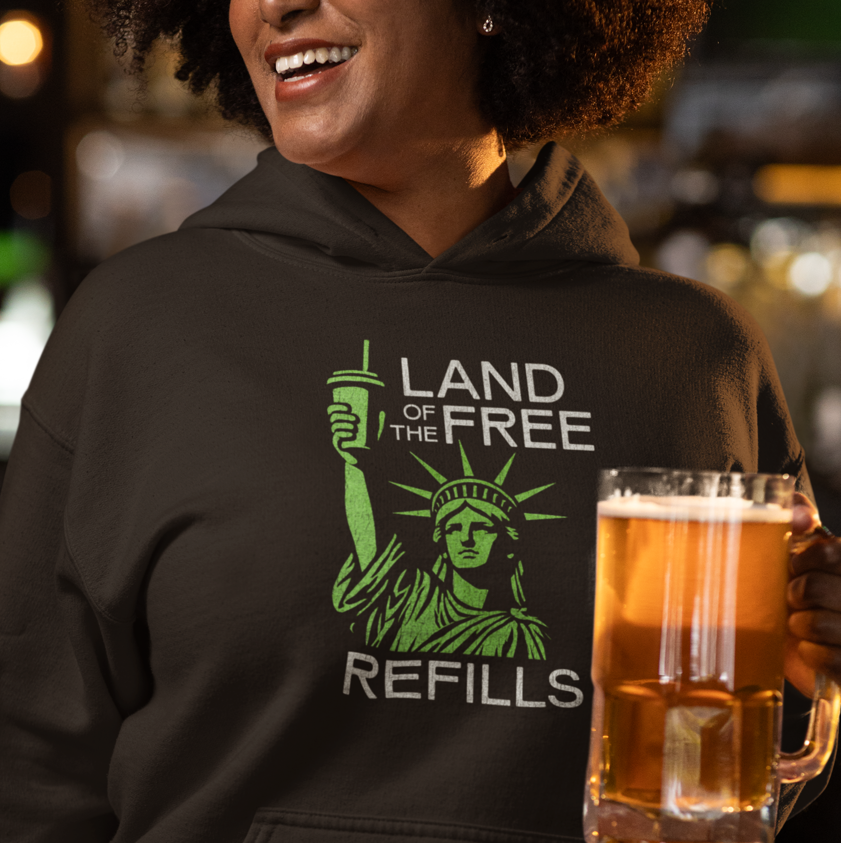 Person wearing a hoodie with 'Land of the Free Refills' design, holding a beer mug in a casual setting.